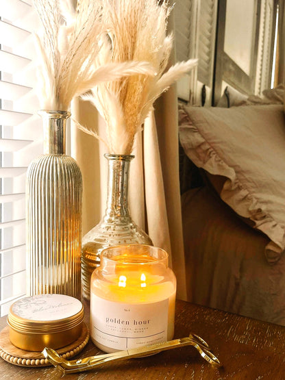 Golden Hour 100% soy wax candle lit on a bedside table, styled with pampas grass in silver vases and a gold wick trimmer. Hand-poured, eco-friendly candle with non-toxic fragrance notes of amber, lemon, ginger, patchouli, and wood, perfect for creating a cozy, warm ambiance in a modern bedroom.